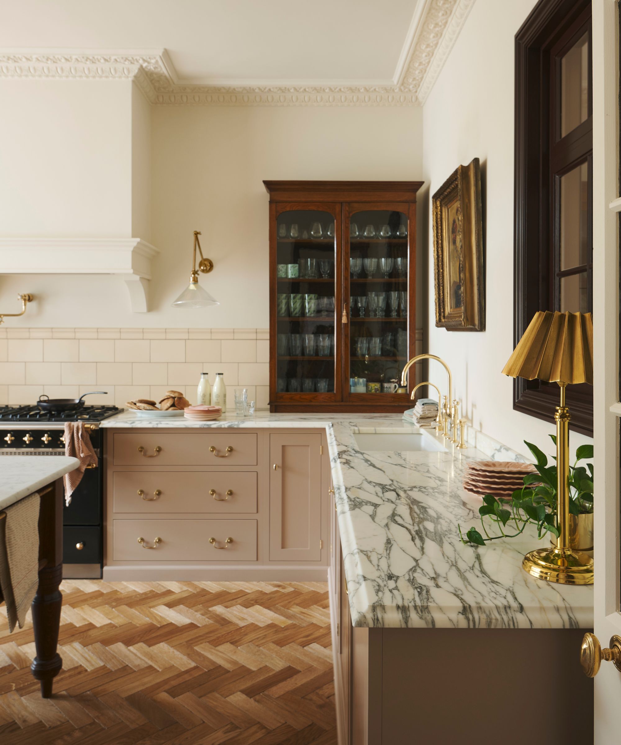 The corner of a Victorian kitchen with pink cabinets, cream walls, and an antique wooden cabinet sat on the marble countertops, storing glassware and mugs
