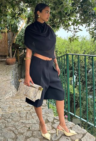 A snakeskin purse is shown in a street-style photo of a woman standing outside wearing gold earrings, a black blouse, black long shorts, gold bangles, yellow slingback heels, and a python clutch bag