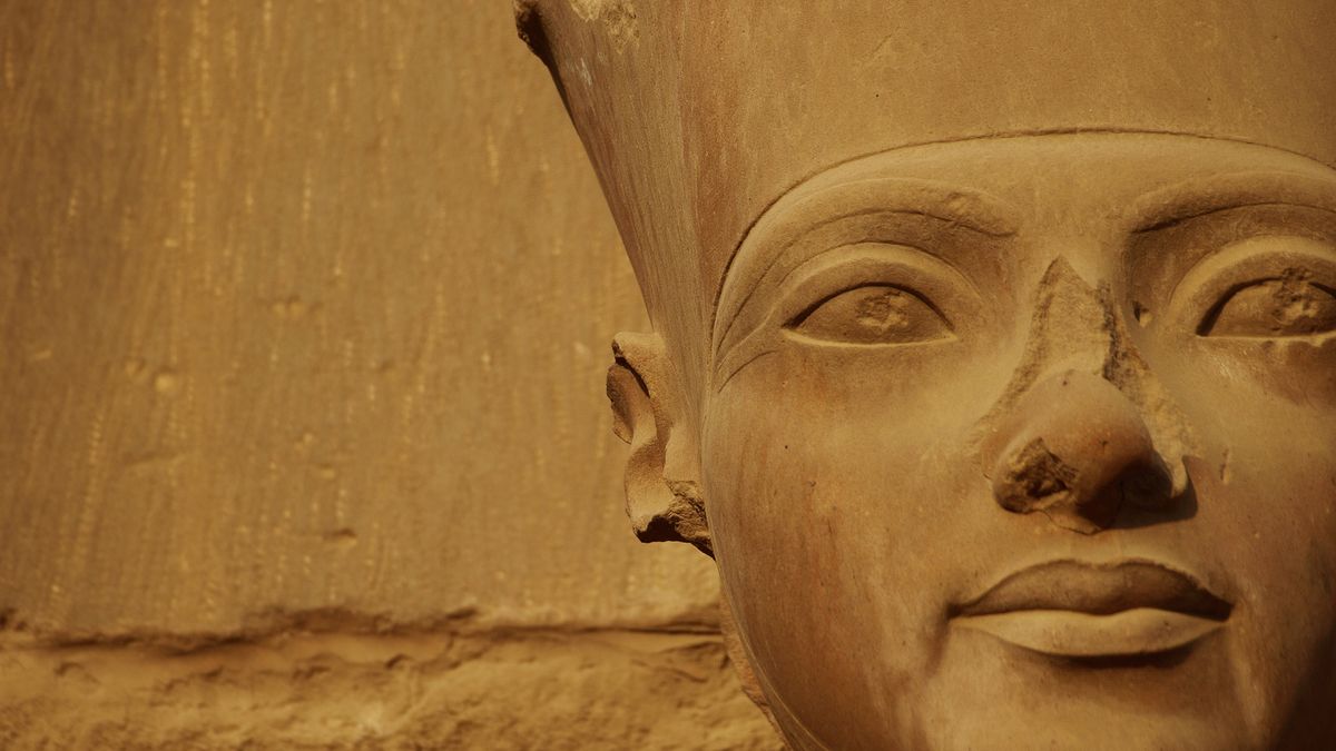 We see the closeup of a stone statue depicting a man wearing a hat. The statue and the background are sandy colored.