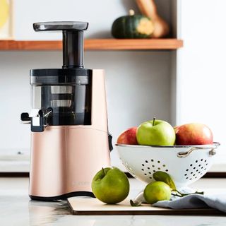 Hurom H-AA slow masticating juicer in rose gold on kitchen counter with colander filled with apples