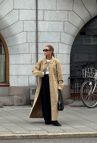 The winter capsule wardrobe item, wool trousers, is shown in a photo of a woman standing outside on a sidewalk wearing a tan suede trench coat over a white t-shirt with black wool trousers, black flats, black sunglasses, and a black bag