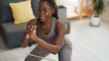 Happy black athletic woman doing squats with resistance band on her around her legs at home.