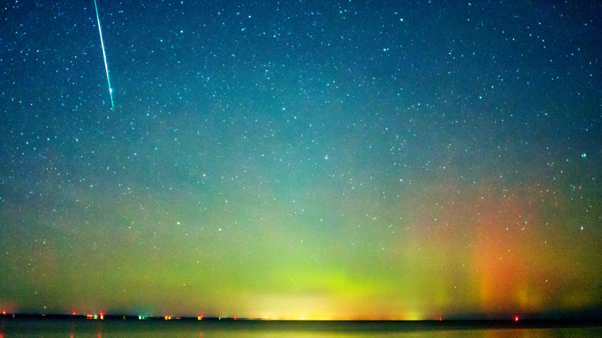 A meteor is photographed streaking Earthward through aurora brightened skies at night above a lake.