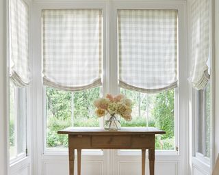 Windows with cream and white gingham Roman shades and a wooden table with vase of pastel colored hydrangea flowers
