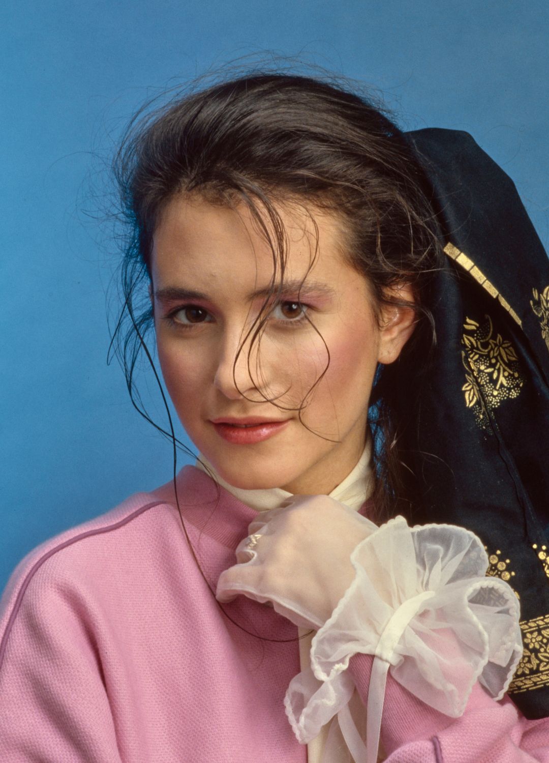 A young woman modelling a pink sweatshirt with a white scarf, wearing a white lace glove on her left hand, during a studio fashion shoot against a blue background for 'Tiger Beat' magazine