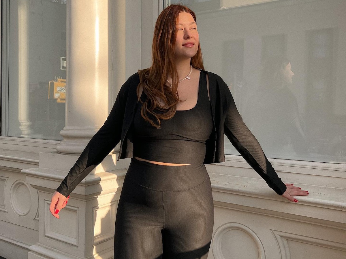 Mid-size fashion influencer Raeann Langas poses against a building in NYC wearing a workout outfit cropped zip-up hoodie, black sports bra, and black leggings