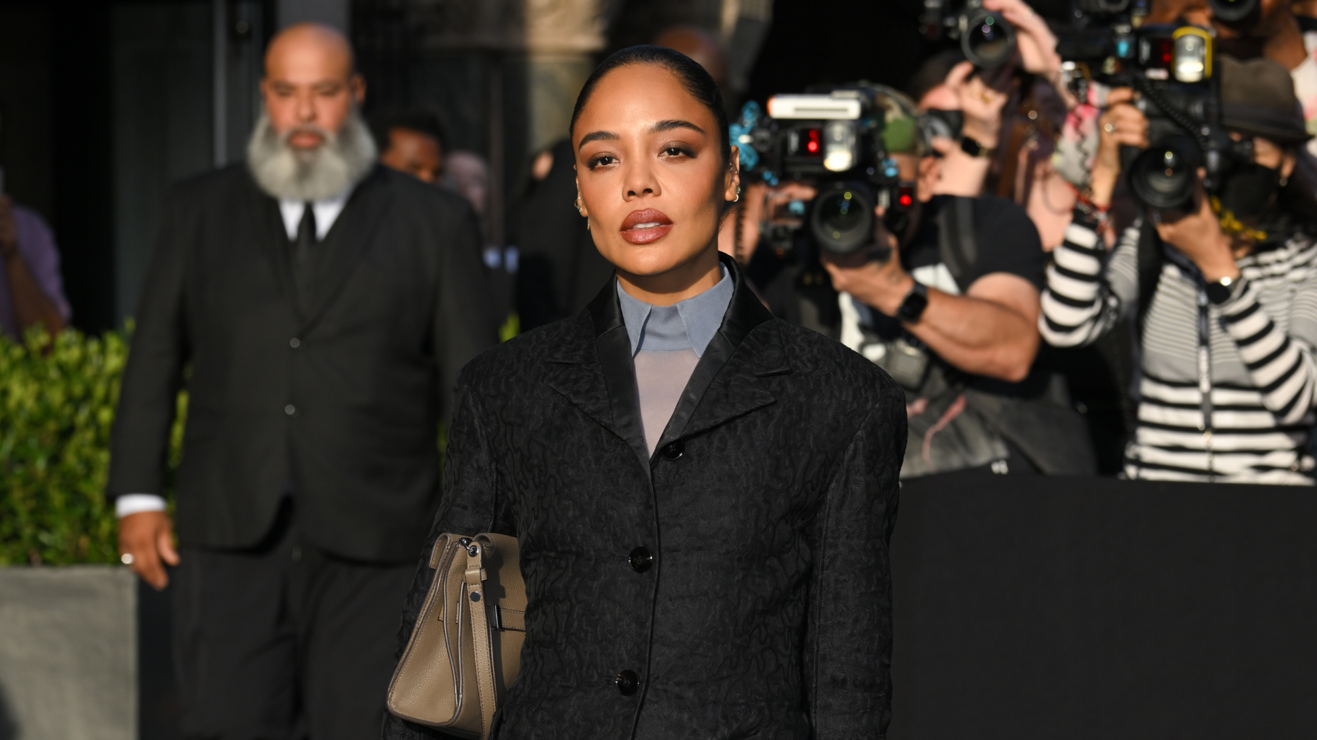 A woman with a sleek bun hairstyle wearing a blazer and collared shirt while holding a taupe colored bag.