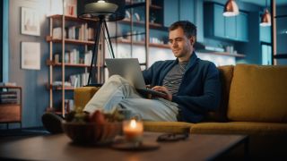 A man sitting on a coach and using a laptop in his lap in a room that looks like a living room