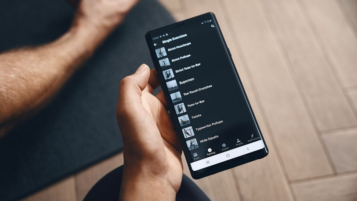 A smartphone showing the Freeletics app held in someone&amp;#039;s hand, with an exercise mat on a wooden floor in the background