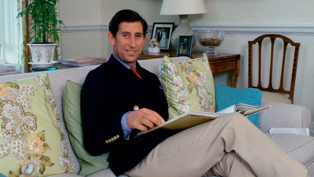 Prince Charles wearing khakis and a black blazer sitting on a sofa with green floral pillows in his living room with a table and lamp behind him