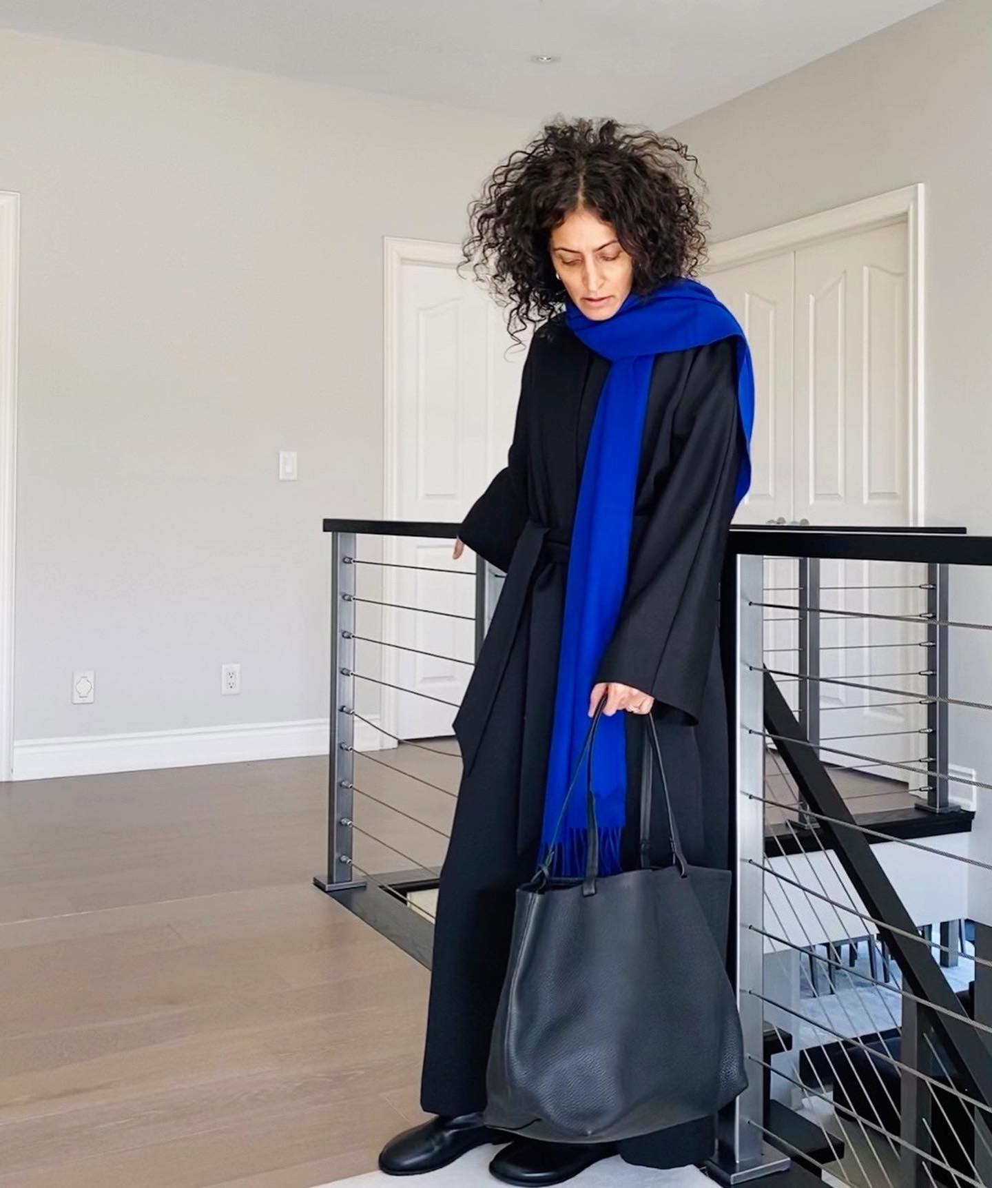 Woman wears black coat, black boots, blue scarf and carries black tote bag