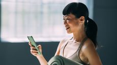 Woman looking at phone while holding a yoga mat under one arm to explore the best Pilates apps