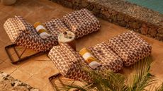 Two patterned outdoor loungers with matching ottomans sit on a terracotta-tiled terrace beside a stone wall overlooking the sea.