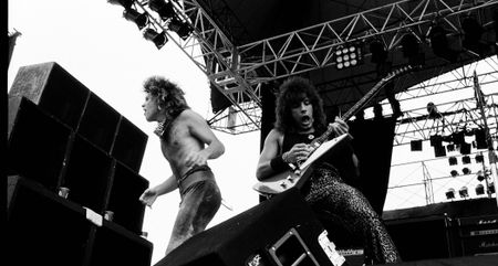 A black and white live shot of Richie Sambora playing his iconic modded Gibson Explorer in 1984, onstage with a shirtless Jon Bon Jovi to his right.