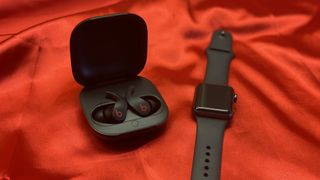 Les écouteurs sans fil Beats Fit Pro dans leur boîtier de chargement sur un fond rouge à côté d'une Apple Watch.