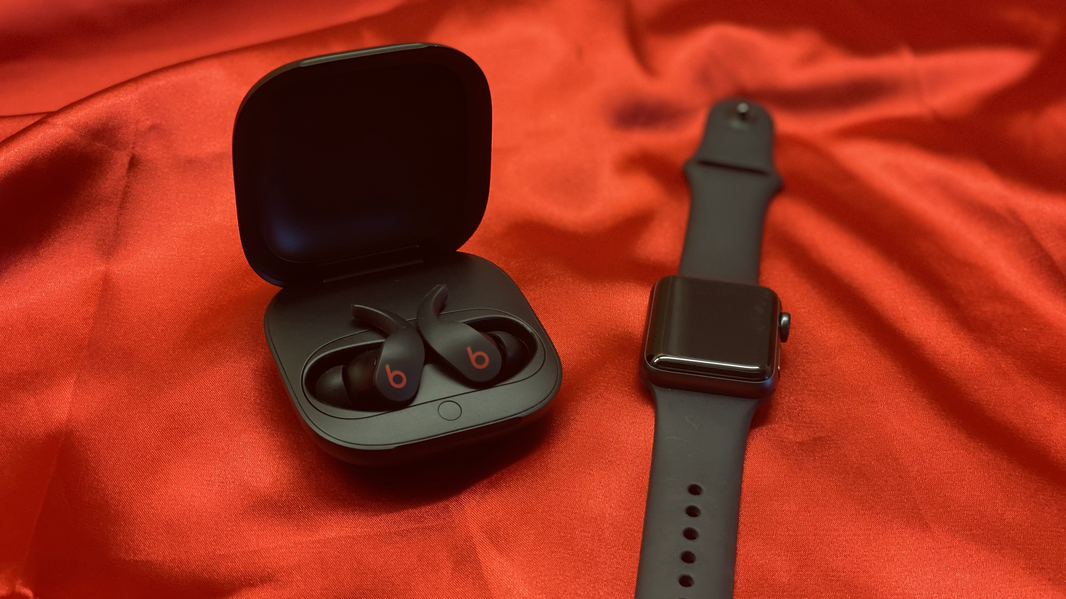 The Beats Fit Pro earbuds in their charging case on a red backdrop next to an Apple Watch
