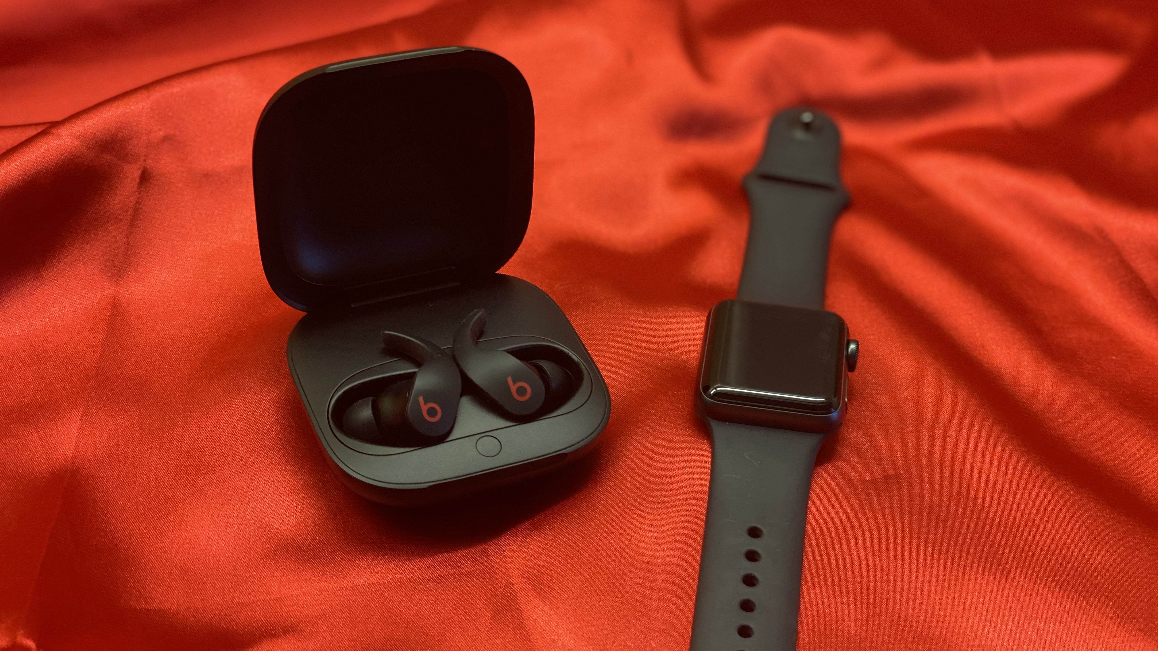 Beats Fit Pro-öronsnäckorna i deras laddningsfodral på en röd bakgrund bredvid en Apple Watch