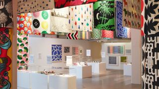 An exhibition space displays various colorful and patterned textiles hanging from the ceiling, with framed prints and display cases featuring designs on pedestals below.