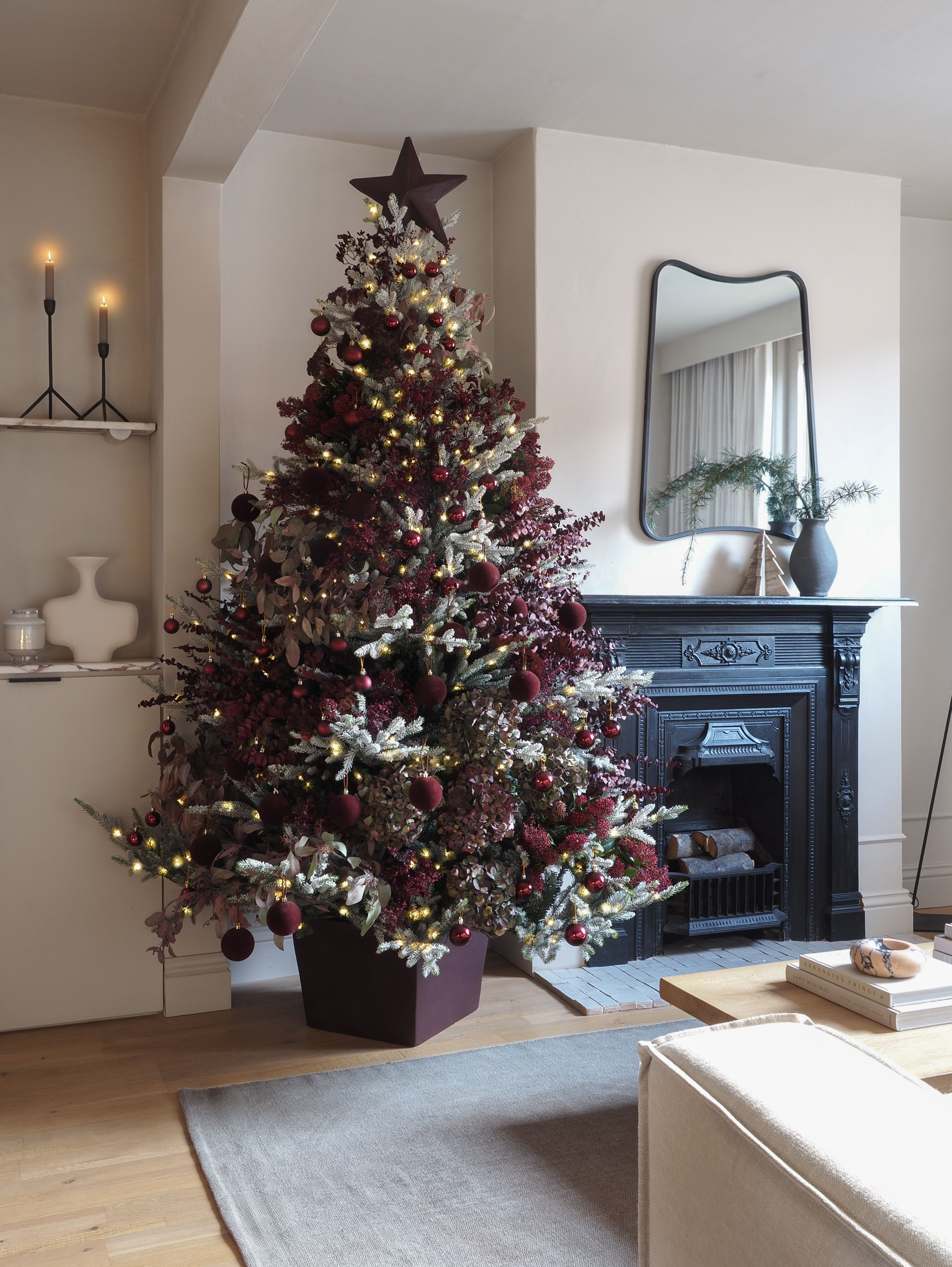 a burgundy christmas tree in a neutral living room next to an ornate fireplace with a sculptural mirror hanging above, and shelves on the side decorated with vases and candles