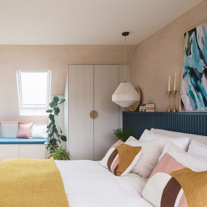 A colourful bedroom with dormer windows and pale pink walls. The double bed is dressed in white bedding with yellow, pink and burgundy accents.