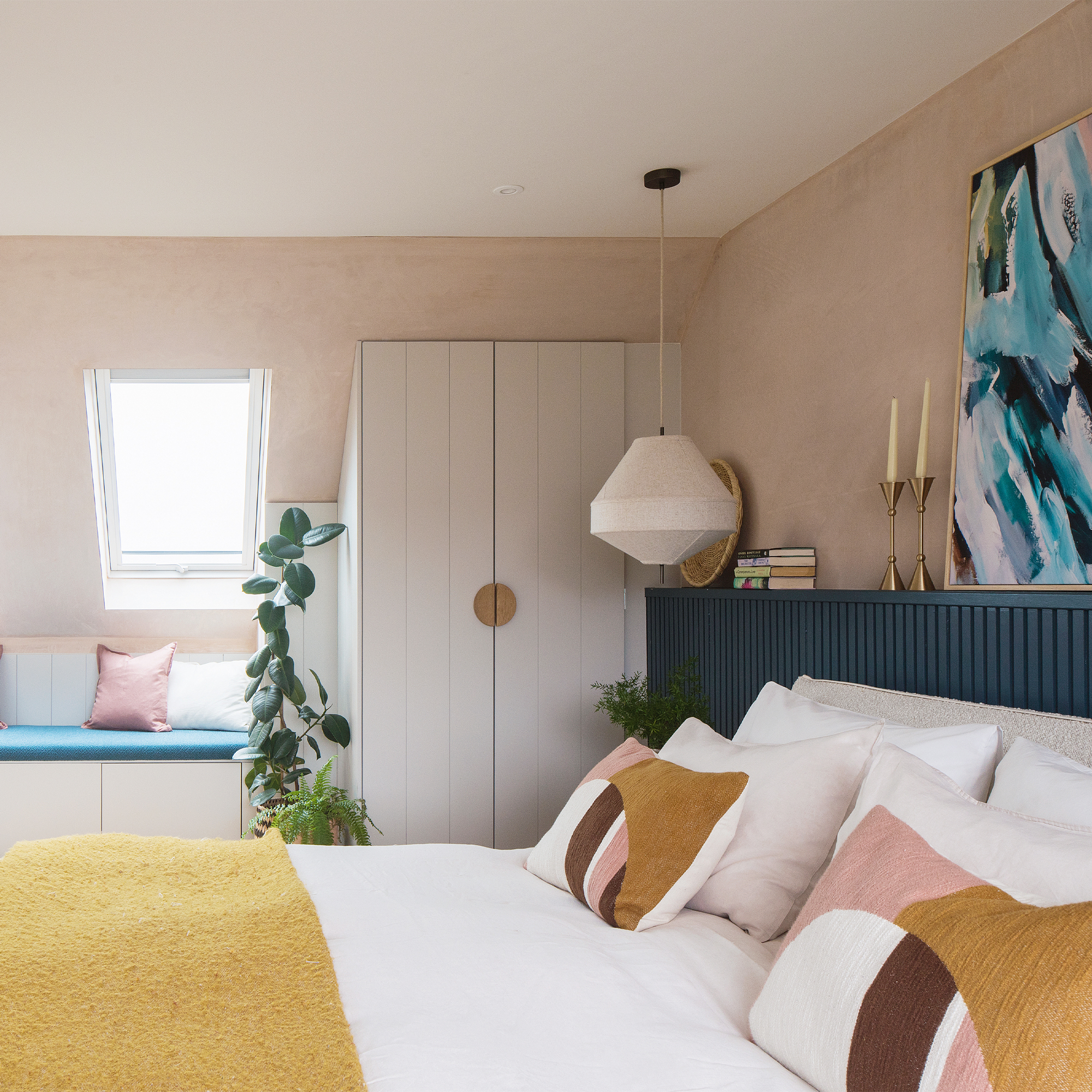 A colourful bedroom with dormer windows and pale pink walls. The double bed is dressed in white bedding with yellow, pink and burgundy accents.