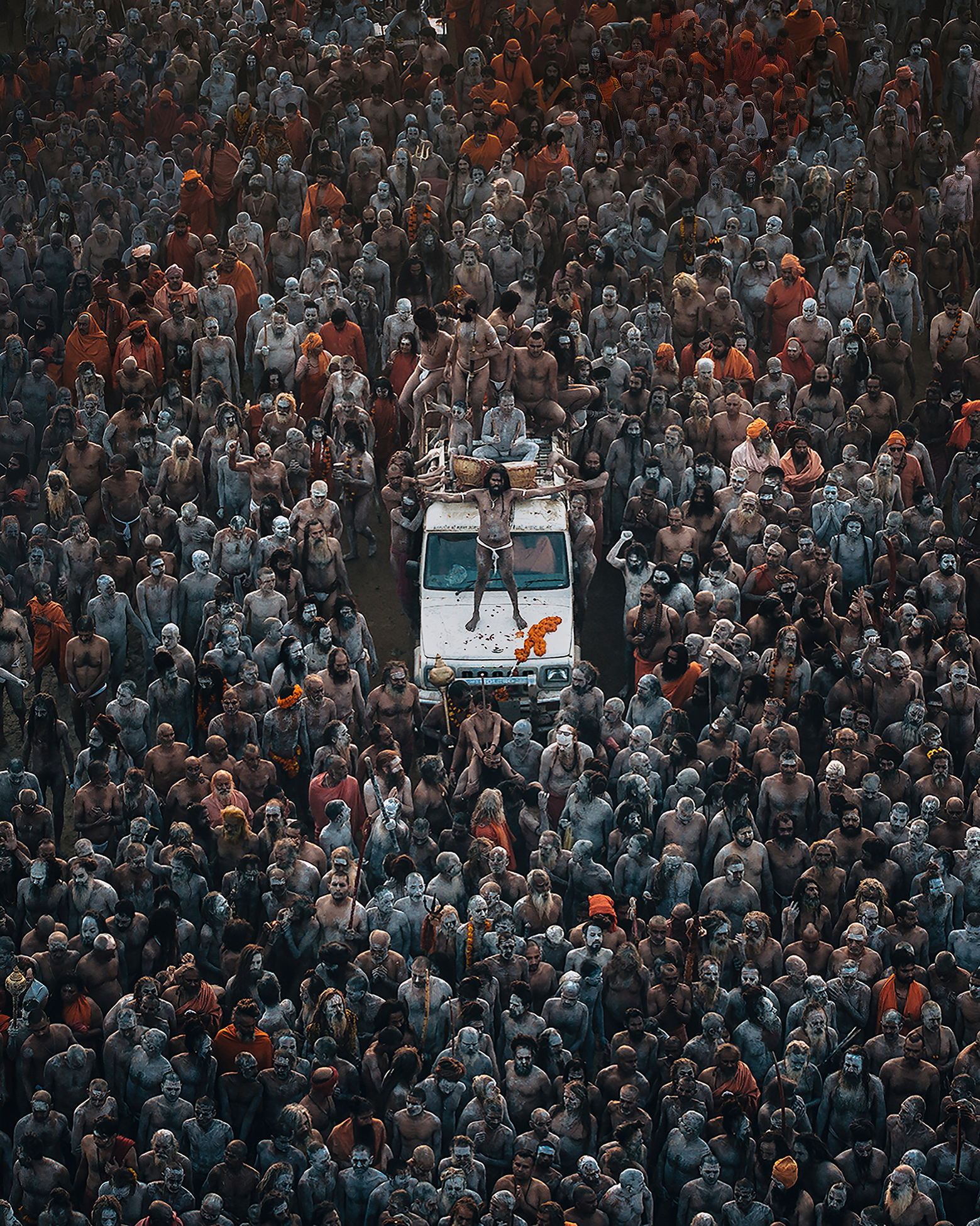 Aerial view of a large congregation of people covered in ash and paint, grouped around a vehicle, engaged in a vibrant spiritual gathering