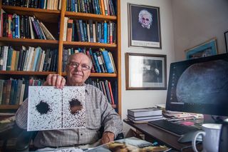 a man sits in an office with a bookshelf behind him, he is holding up two images.