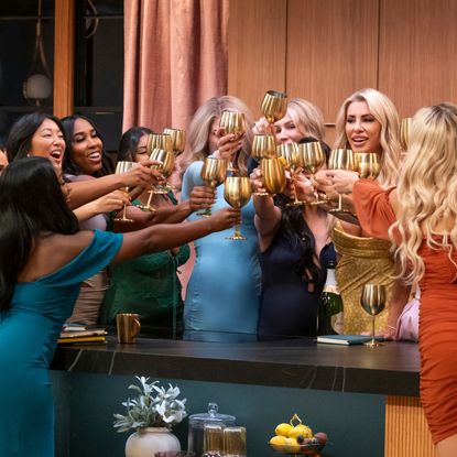 A group of women (l-r: Tyler Lanier, Dynasty Ballard, Emma Betsinger, Bry Thomas, Priyanka Grandhi, Amber Morrison, Christine Hamilton, Elissa Finley, and Rosalyn Ransaw) toast with gold wine glasses as they stand over a kitchen island, in the 'Love is Blind' season 10 premiere.