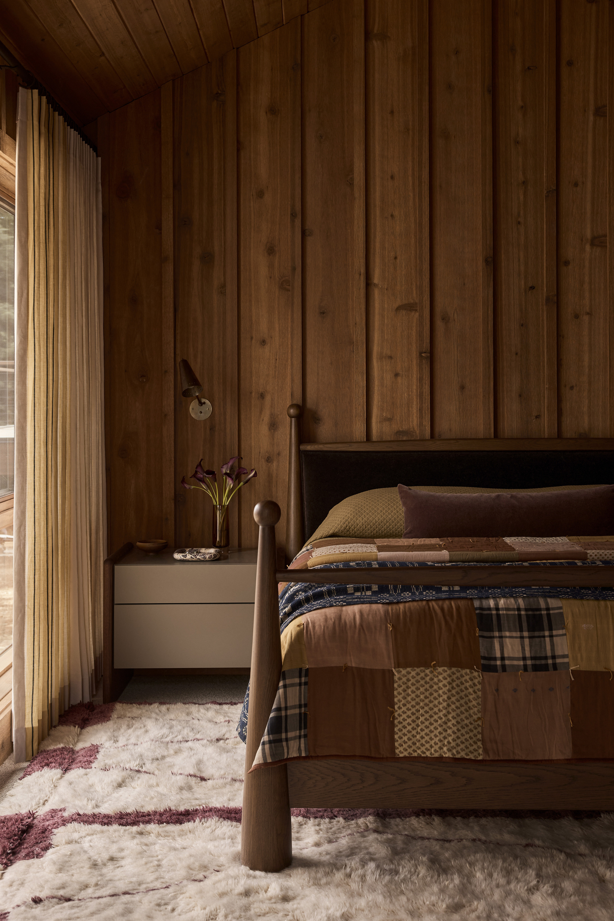 Image of a wooden bed with a patchwork quilt on it in a room that has wooden paneled walls. There is a white and red shag rug on the ground.