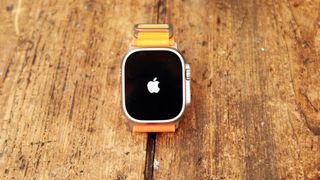 En Apple Watch Ultra med ett orange armband ligger på ett träbord utomhus och visar Apple-logon på skärmen.