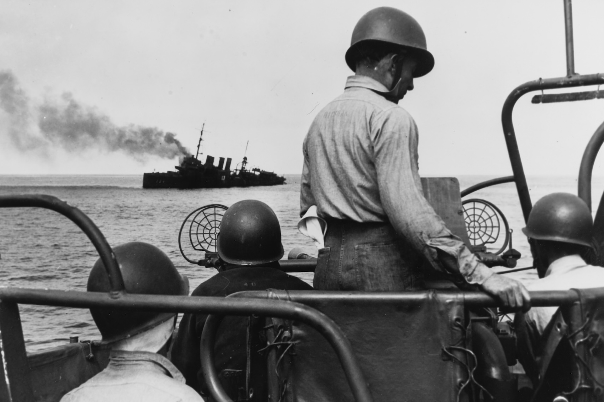 Uma foto em preto e branco de um grupo de marinheiros em um barco olhando para um navio afundando no horizonte