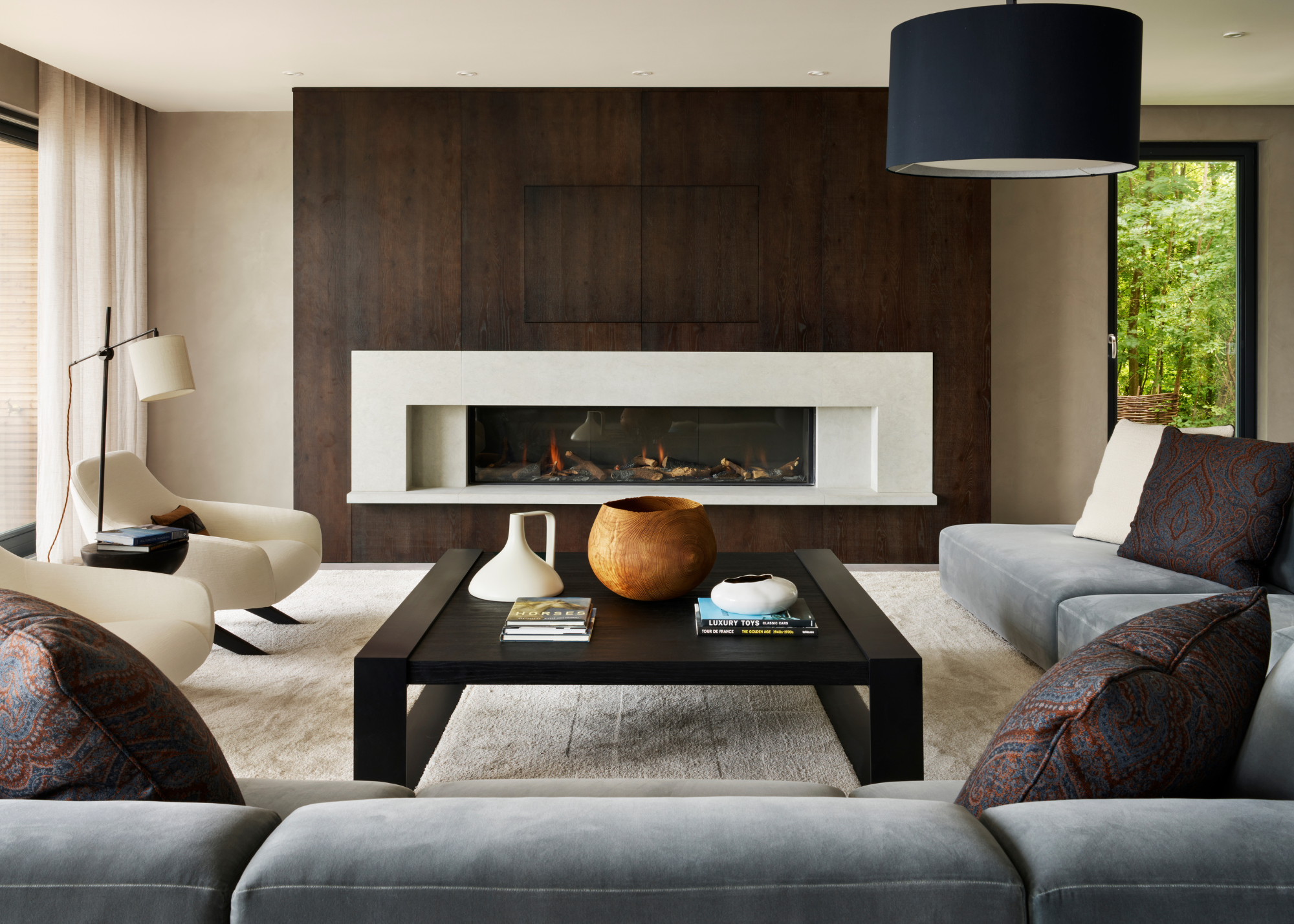A modern living room with a large fireplace, l-shaped sofa and a large dark coffee table in the center with large decorative objects and books styled on top