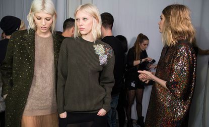 Room full of people and three female models wearing looks from No 21's collection. One model is wearing a light brown jumper, semi-sheer peach skirt and dark green embellished coat. Next to her is a model wearing a dark green jumper with embellishment by the shoulder and a black semi-sheer skirt. The last model is wearing a multicoloured sequin piece