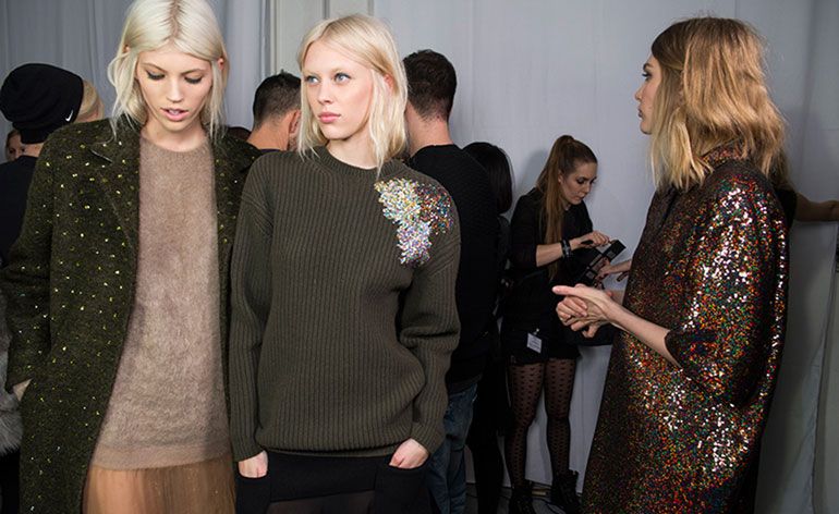 Room full of people and three female models wearing looks from No 21&#039;s collection. One model is wearing a light brown jumper, semi-sheer peach skirt and dark green embellished coat. Next to her is a model wearing a dark green jumper with embellishment by the shoulder and a black semi-sheer skirt. The last model is wearing a multicoloured sequin piece