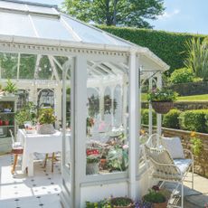 Ornate conservatory exterior, with dining table inside, metal bench outside, with a lush garden in the background