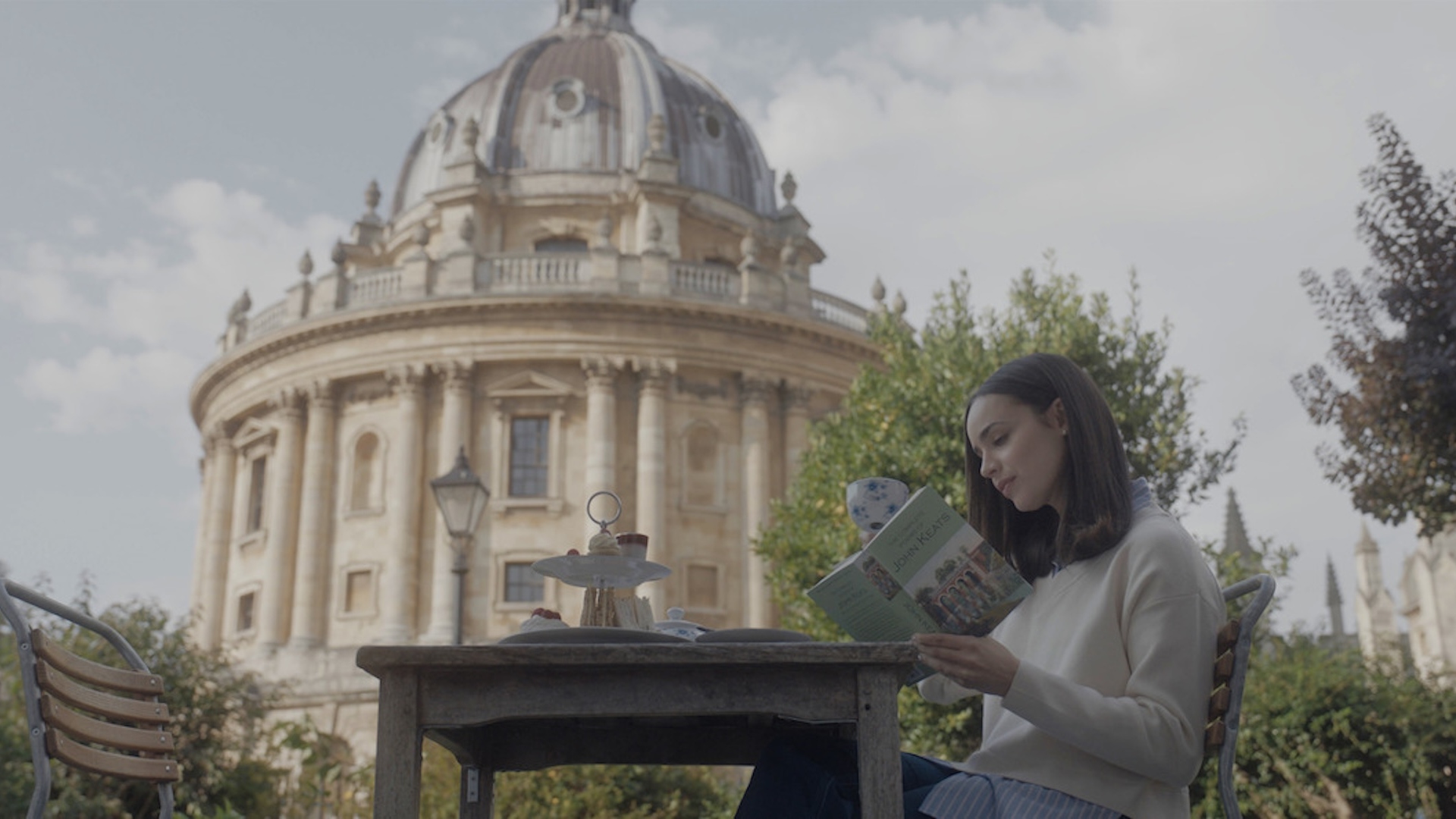 An image of Sofia Carson playing Anna in the film My Oxford Year. She is sitting in front of Oxford's Radcliffe Camera reading a book and drinking tea.
