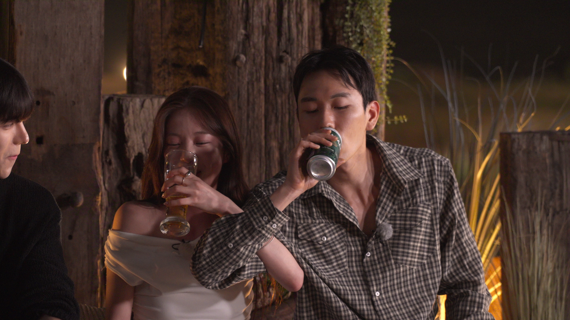 A woman (Lee Joo-young) and a man (Lee Jae-jin) take a love shot while sitting in front of a wooden structure. The woman wears a white blouse and large pendant necklace while drinking from a glass, while the man wears a plaid shirt and drinks from a can. A still from 'Single's Inferno' season 5.