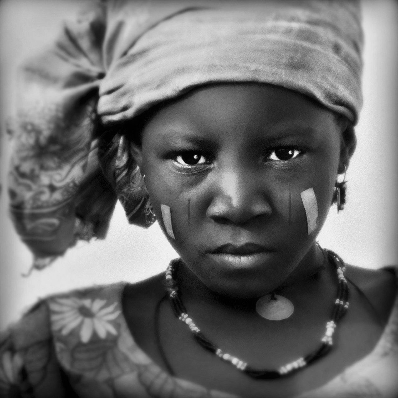 Black and white image showing a black girl wears a patterned headscarf and a necklace, with her upper body visible against a soft background. The image is in black and white