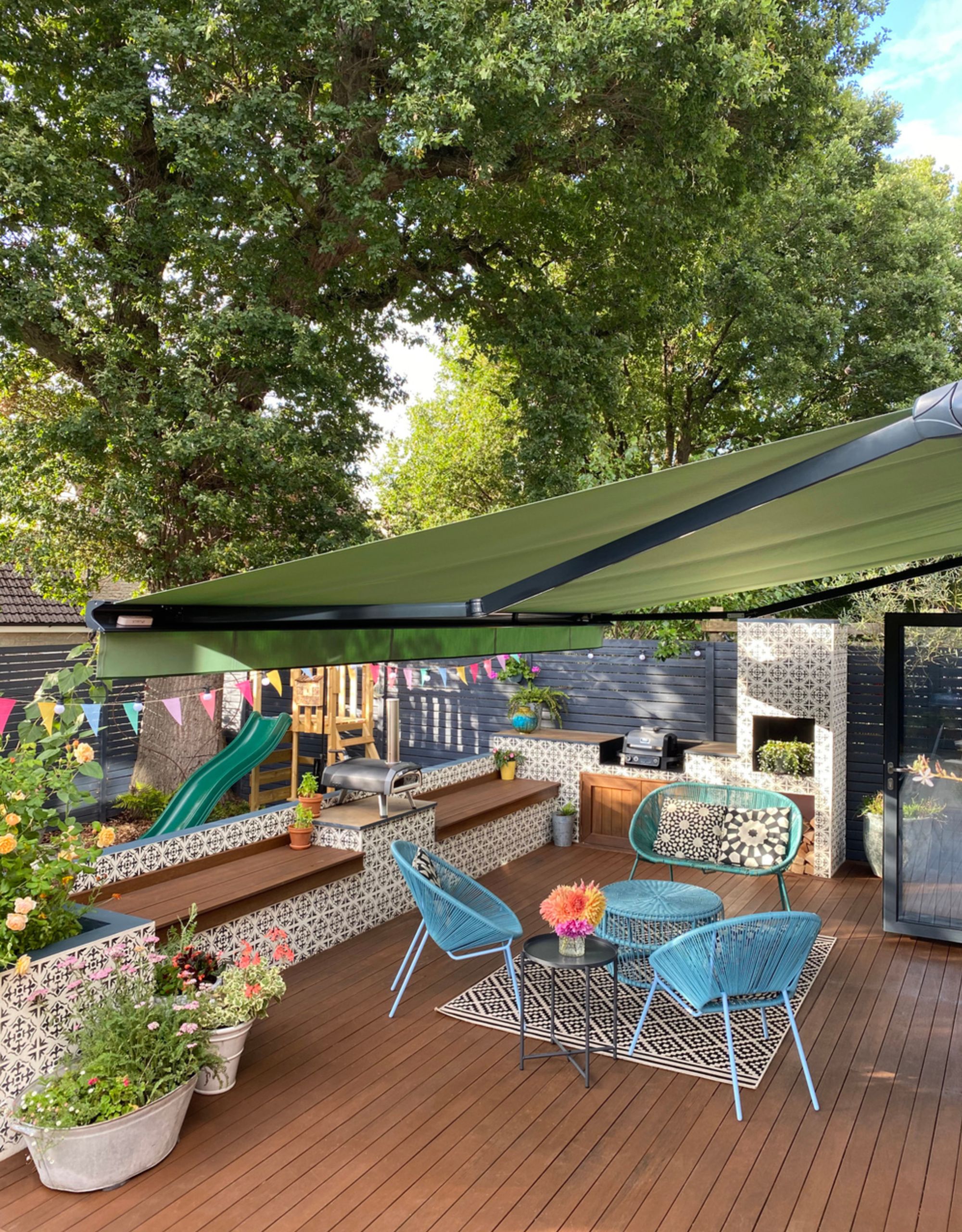 A decking area with a green awning offering shade for an outdoor seating area with a children's play area in the lower area of the garden