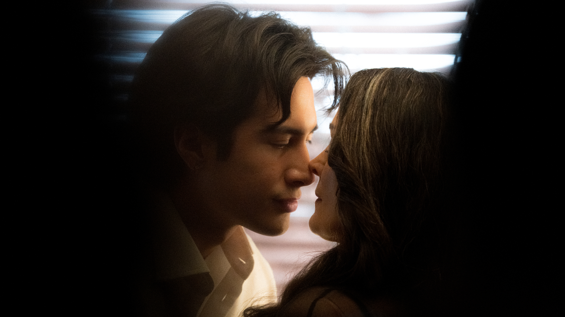 Hudson Williams and Carrie-Anne Moss stand closely together in front of window blinds, seen through shadows, in the Crave tv show 'Yaga.'