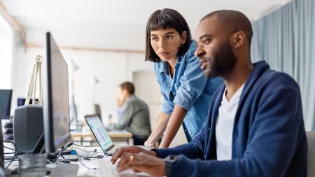 A female office worker looking on as a male office worker types on a keyboard in front of a monitor
