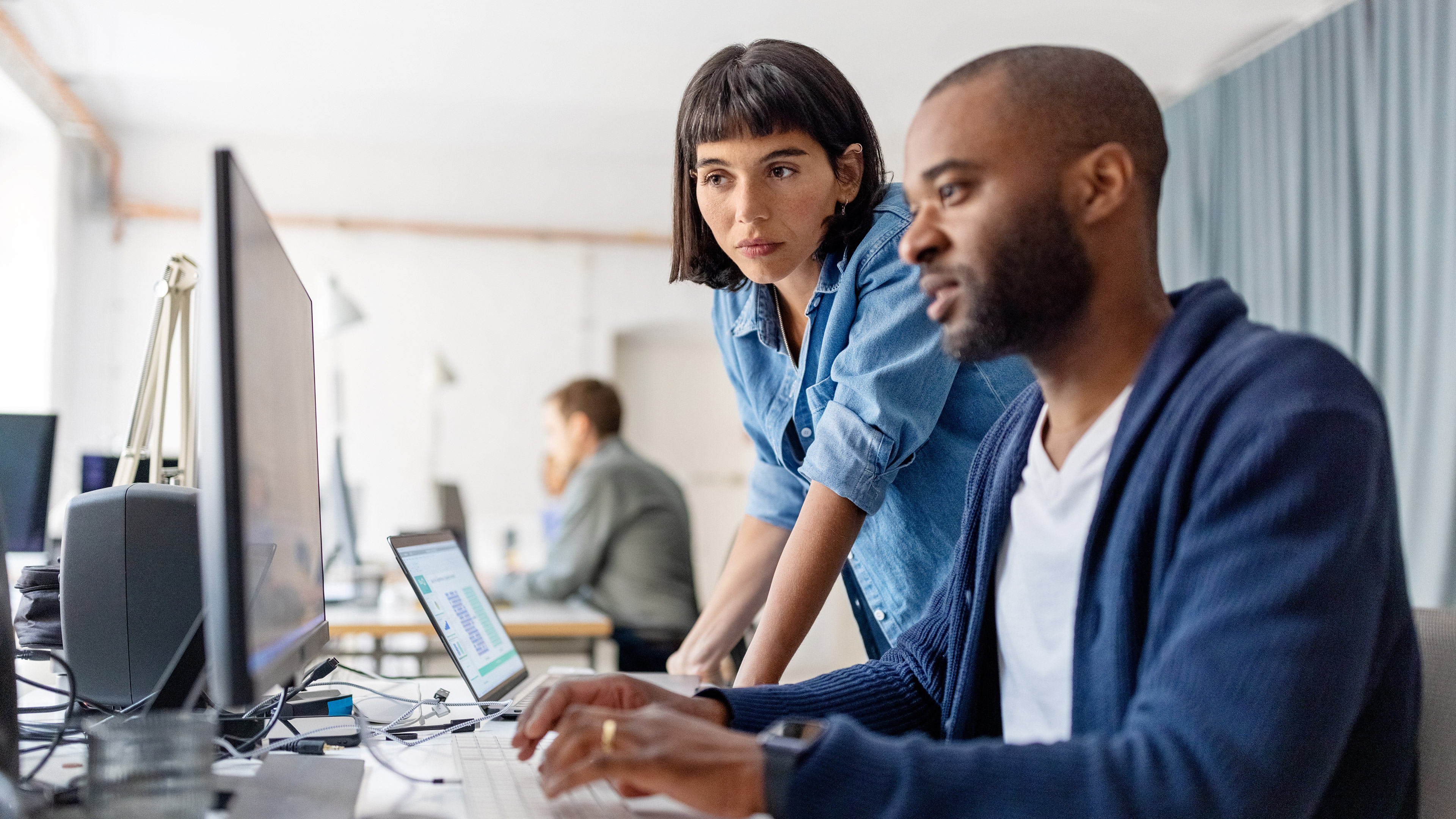A female office worker looking on as a male office worker types on a keyboard in front of a monitor