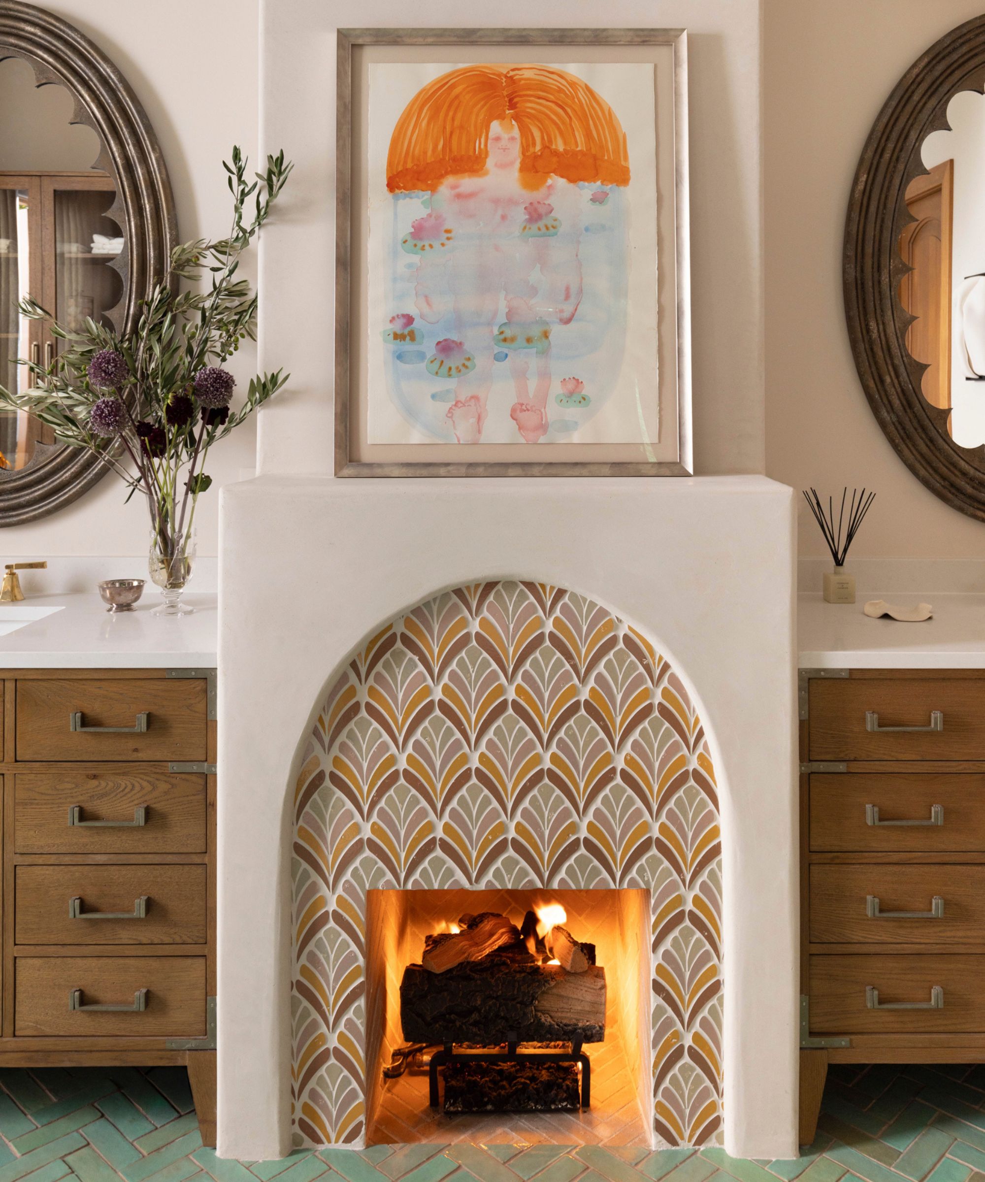 A plaster fireplace with orange and brow tiles and a painting on top. Either side is a wooden vanity topped with a round vanity mirror