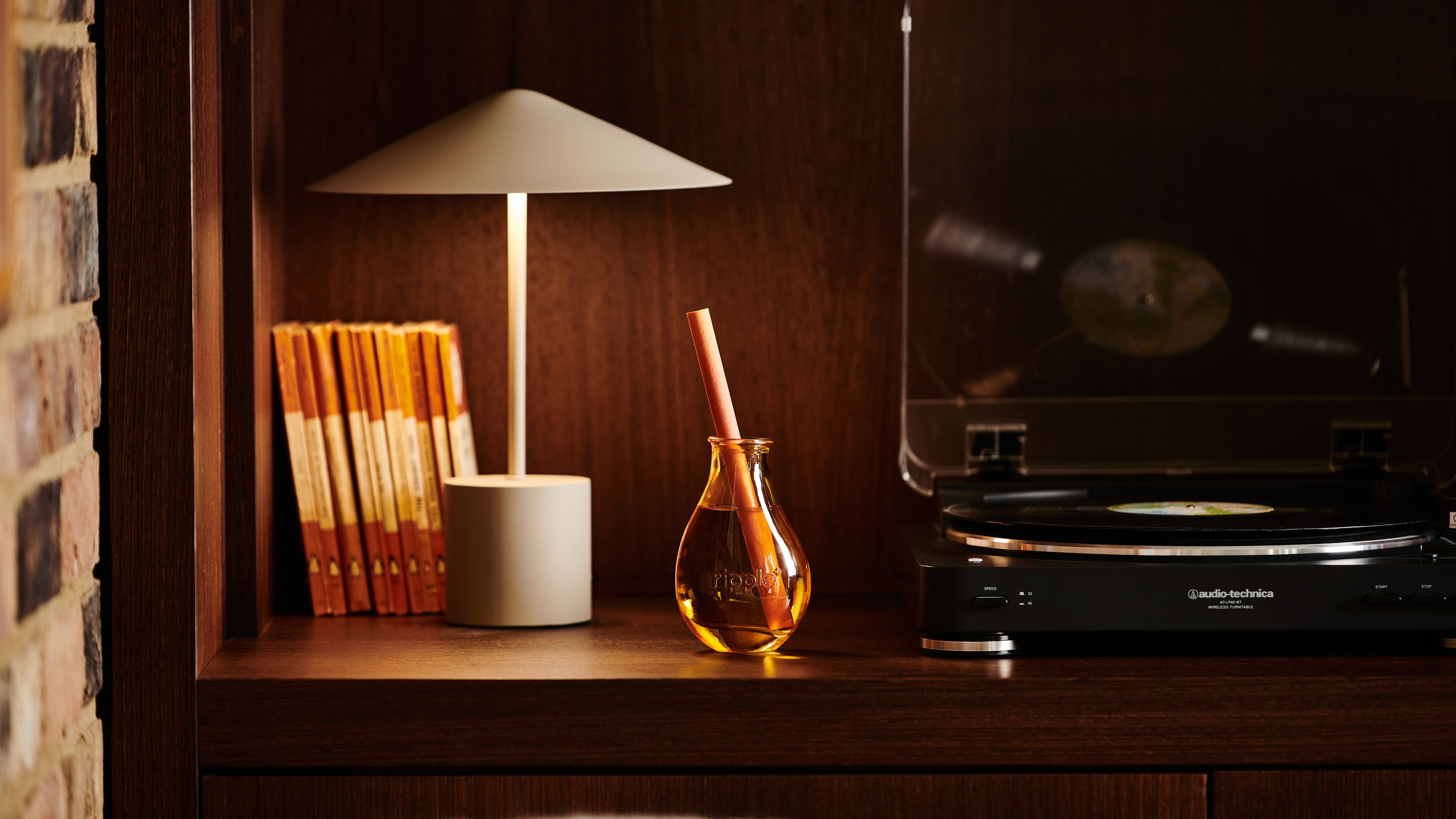 ripple+ Home reed diffuser sitting on a dark wooden bookshelf alongside a sleek white table lamp, books, and a vintage-style record player.