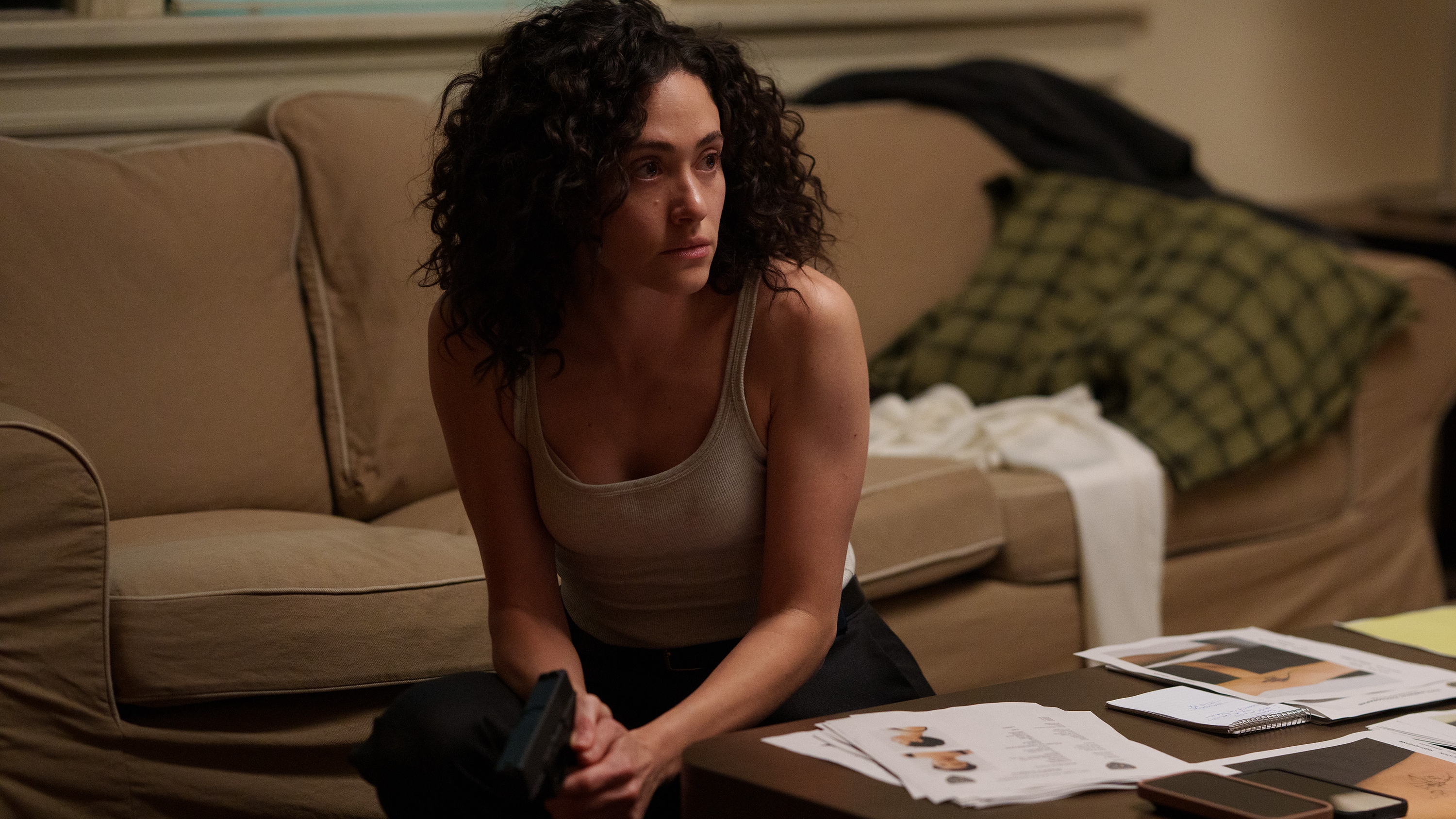 Alice Black (Emmy Rossum) looking alert and holding a gun while crouching down next to a sofa.