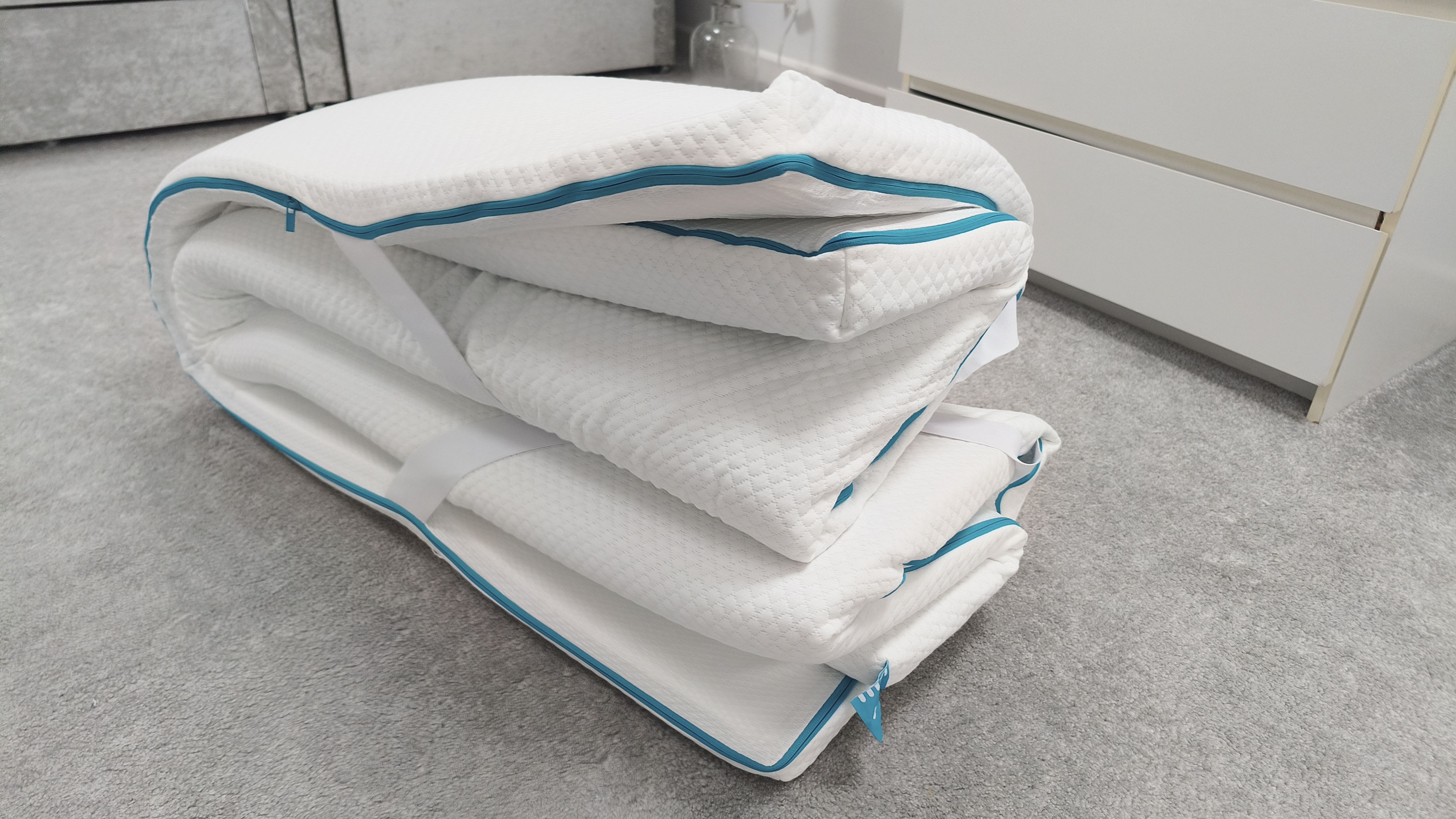 The Simba Cool Foam Mattress Topper folded on a bedroom floor, showing the teal zip detailing and the compact form it takes when not in use.