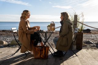 Rosaline (Eve Best) and Cathy (Stockard Channing) sit at a table at a beachfront cafe with take-away coffees
