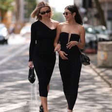 Two women dressed in all black looking at each other, holding handbag, walking towards the camera, one in loafers one in ballet pumps