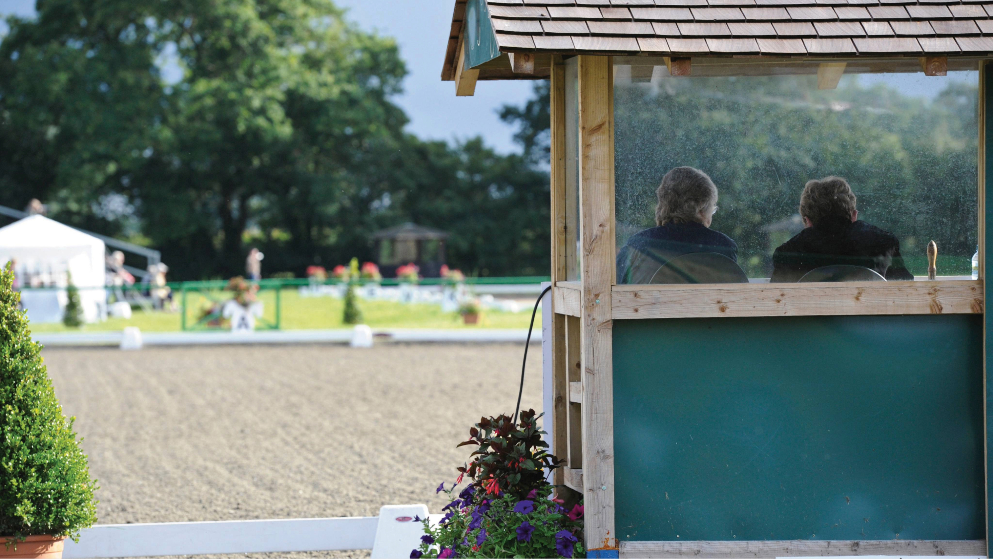 Two dressage judges seen from behind, sitting in a wooden judges&rsquo; box with a glass front and looking out over an empty arena
