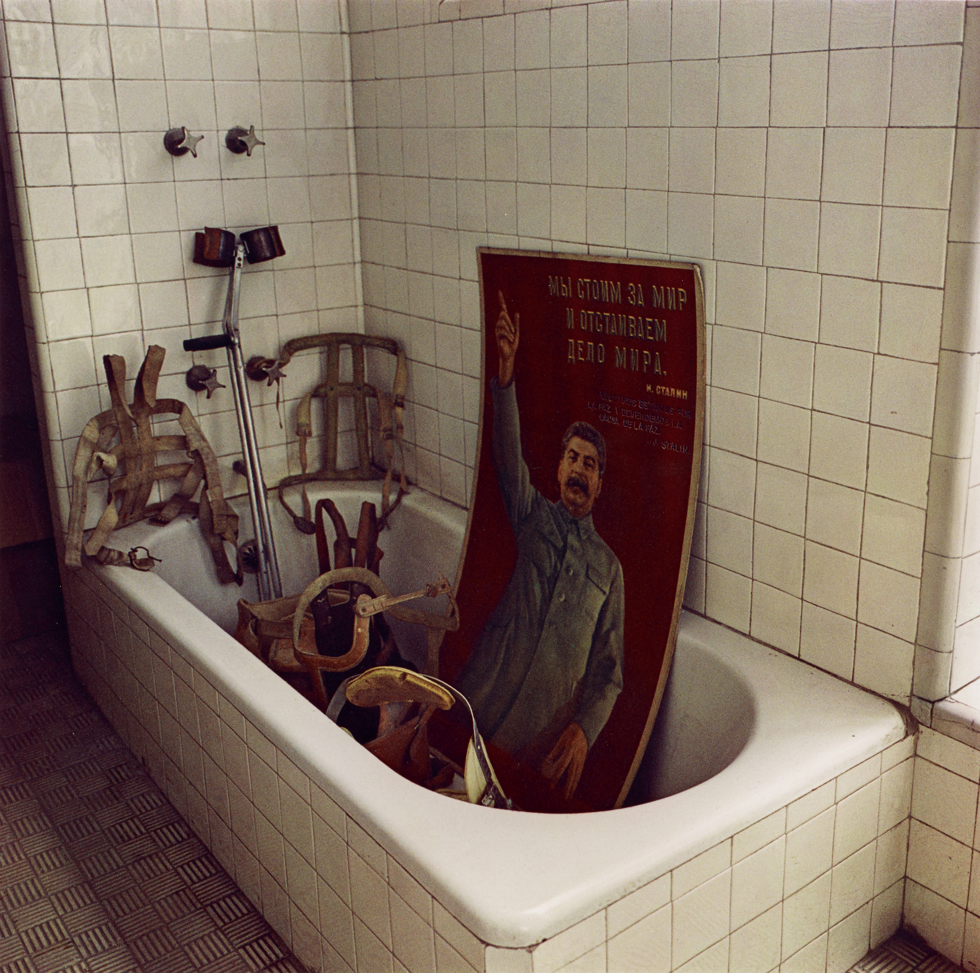 Colour photograph of a white tiled bathtub filled with leather orthopaedic braces, crutches and a faded red Soviet propaganda poster of Stalin.